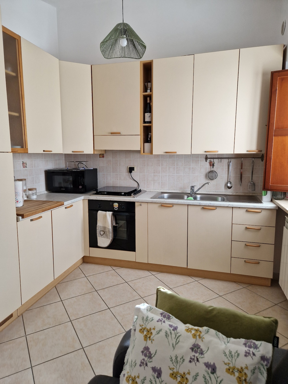 A modern kitchen displays light-colored cabinets and a combination of built-in appliances, including a microwave and an oven. A sink is positioned next to a stove, and a small wooden countertop allows for food preparation. Natural light brightens the space, reflected in the tiled flooring.