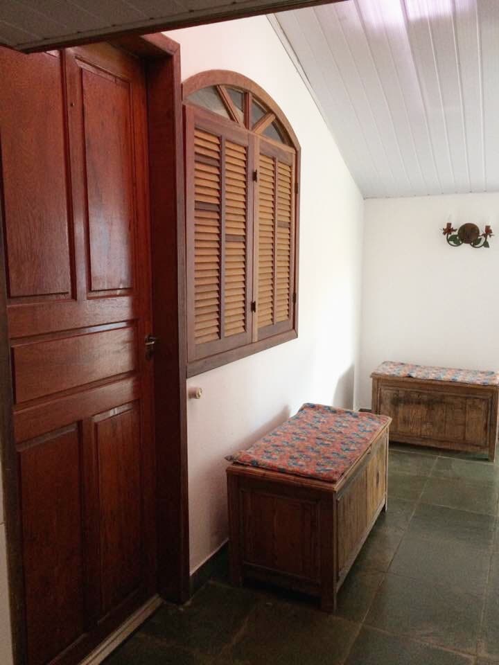The entrance area features two wooden benches adorned with colorful cushions, providing seating. A window with wooden shutters allows natural light to enter, while a wall sconce adds a touch of warmth. The flooring consists of dark stone tiles, enhancing the overall ambiance.