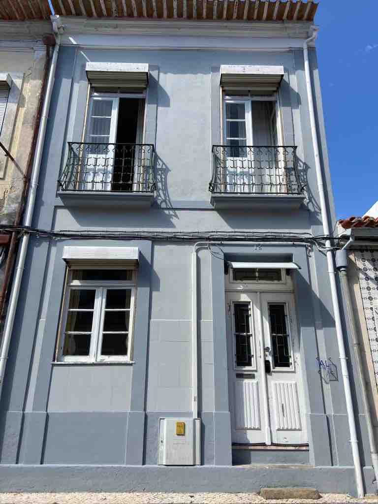A two-story building is shown with a light gray exterior. Two ornate balconies are positioned above the entrance, which features double doors framed by tall windows. The windows are adorned with shutters, adding character to the inviting facade.