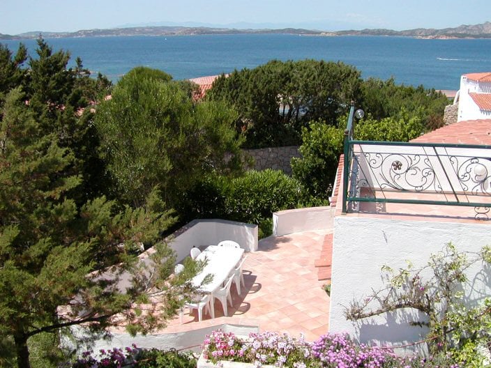 A spacious patio area features a long table surrounded by several chairs, inviting outdoor dining. Lush greenery and blooming flowers line the edges, while views of the sea and distant coastline can be seen in the background under clear skies.