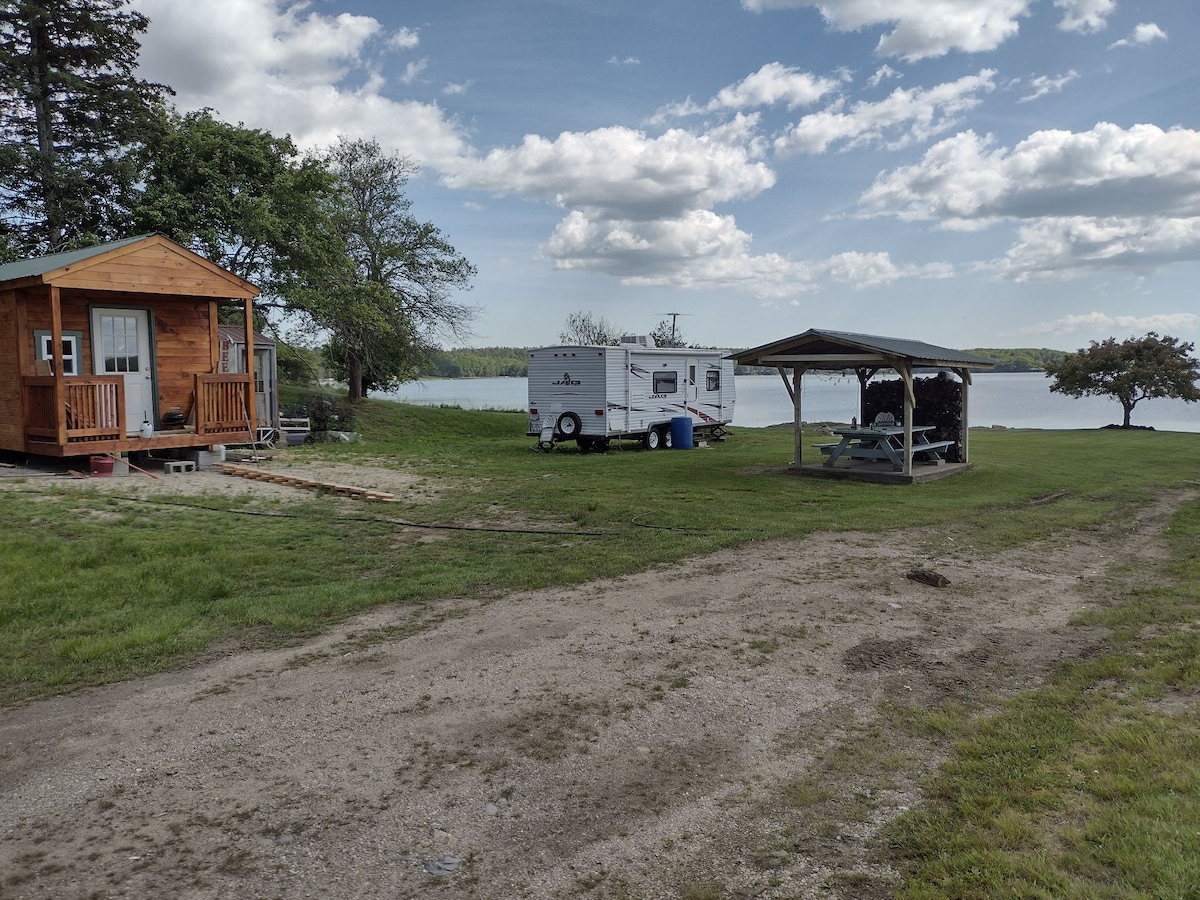 A wooden structure sits alongside a trailer, both positioned on grassy land near the water's edge. A covered pavilion with picnic tables is visible nearby. The scene is set against a backdrop of cloudy blue skies and a serene river, creating a peaceful outdoor environment.