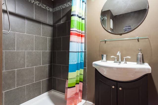 A modern bathroom is presented with sleek grey tile walls and a spacious shower area. A colorful striped shower curtain adds a vibrant touch. A round mirror hangs above a white sink with dark cabinetry below, complemented by simple toiletries and accessories.