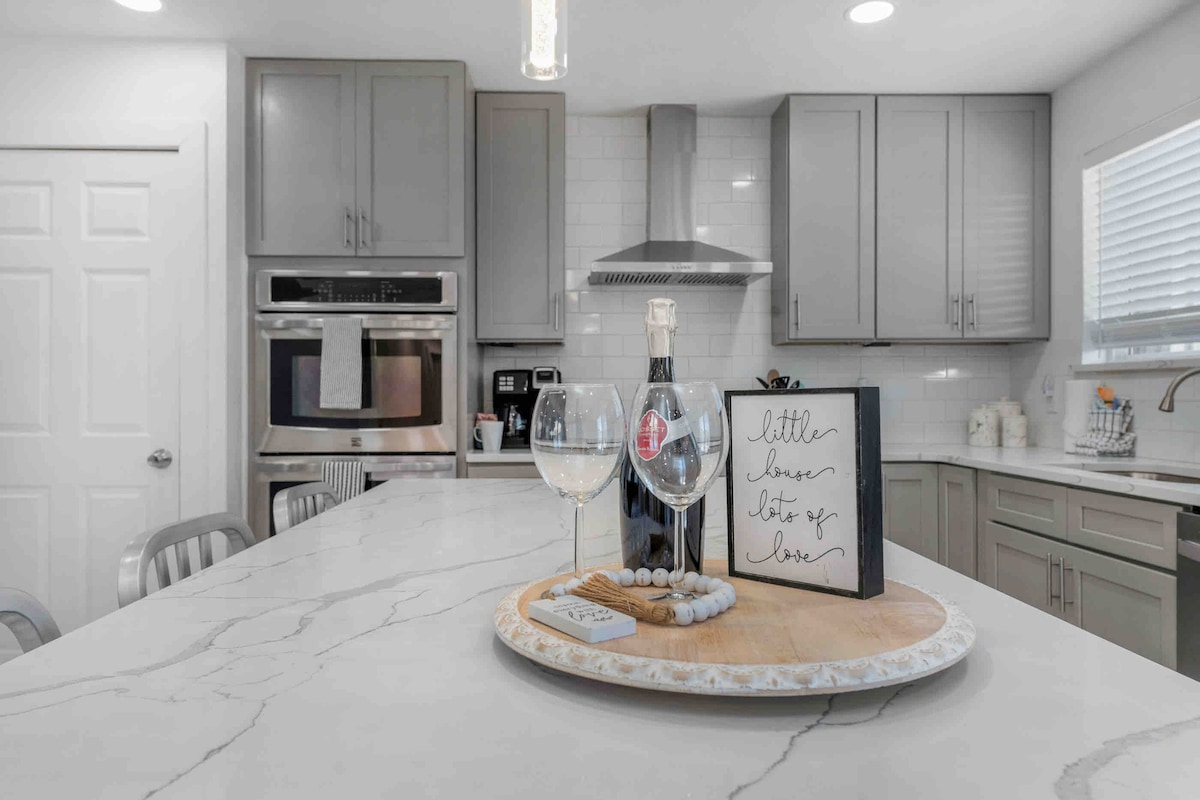 A modern kitchen features sleek gray cabinetry and a spacious marble countertop. On the counter, two glasses of wine rest beside a decorative wooden tray with a handwritten note and small pebbles, adding a touch of charm to the culinary space.