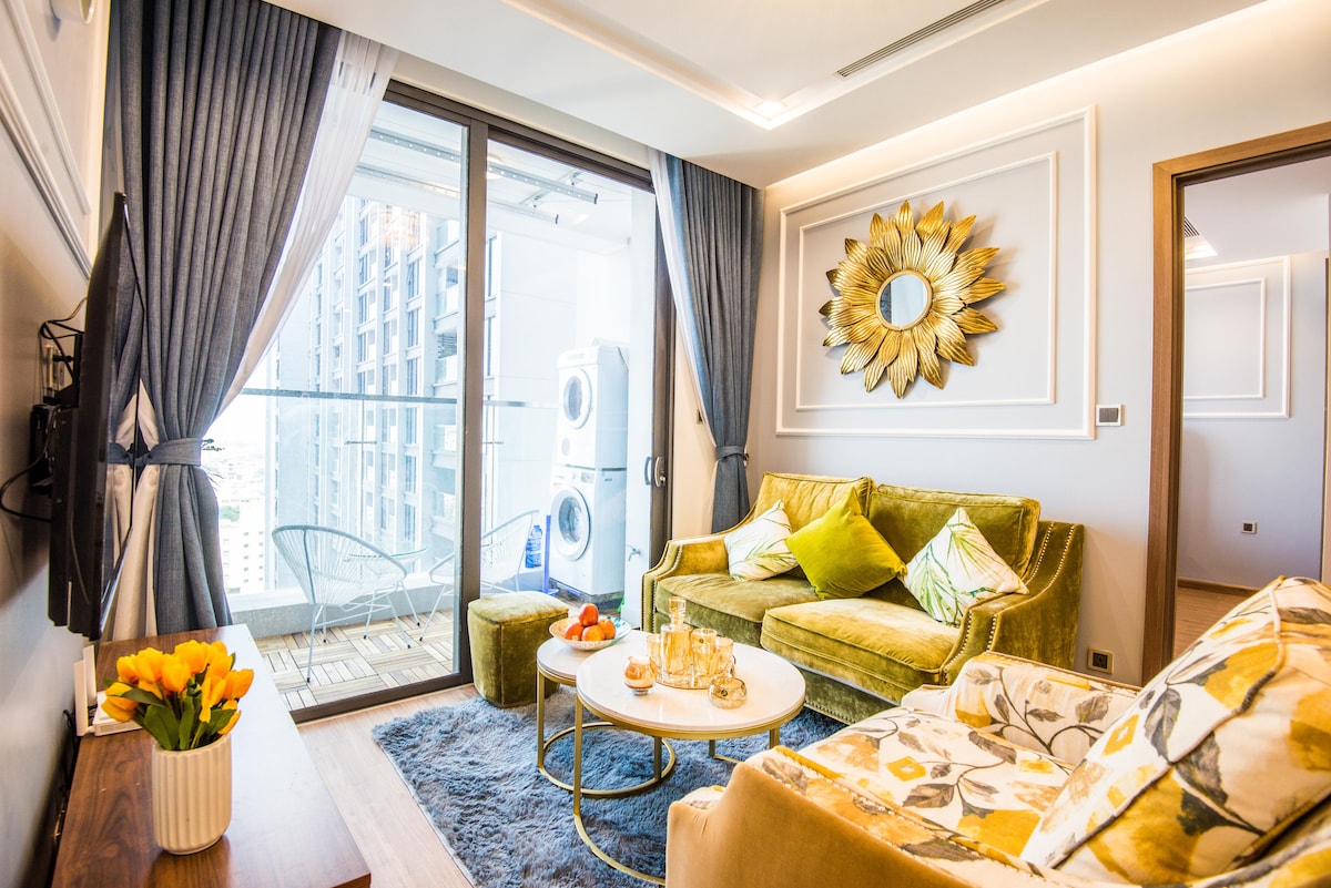 A modern living area features a vibrant green couch adorned with decorative pillows, complemented by a circular coffee table. A large wall mirror with a sunburst design enhances the space, while sliding glass doors lead to a balcony with seating. A washing machine is visible on the balcony.