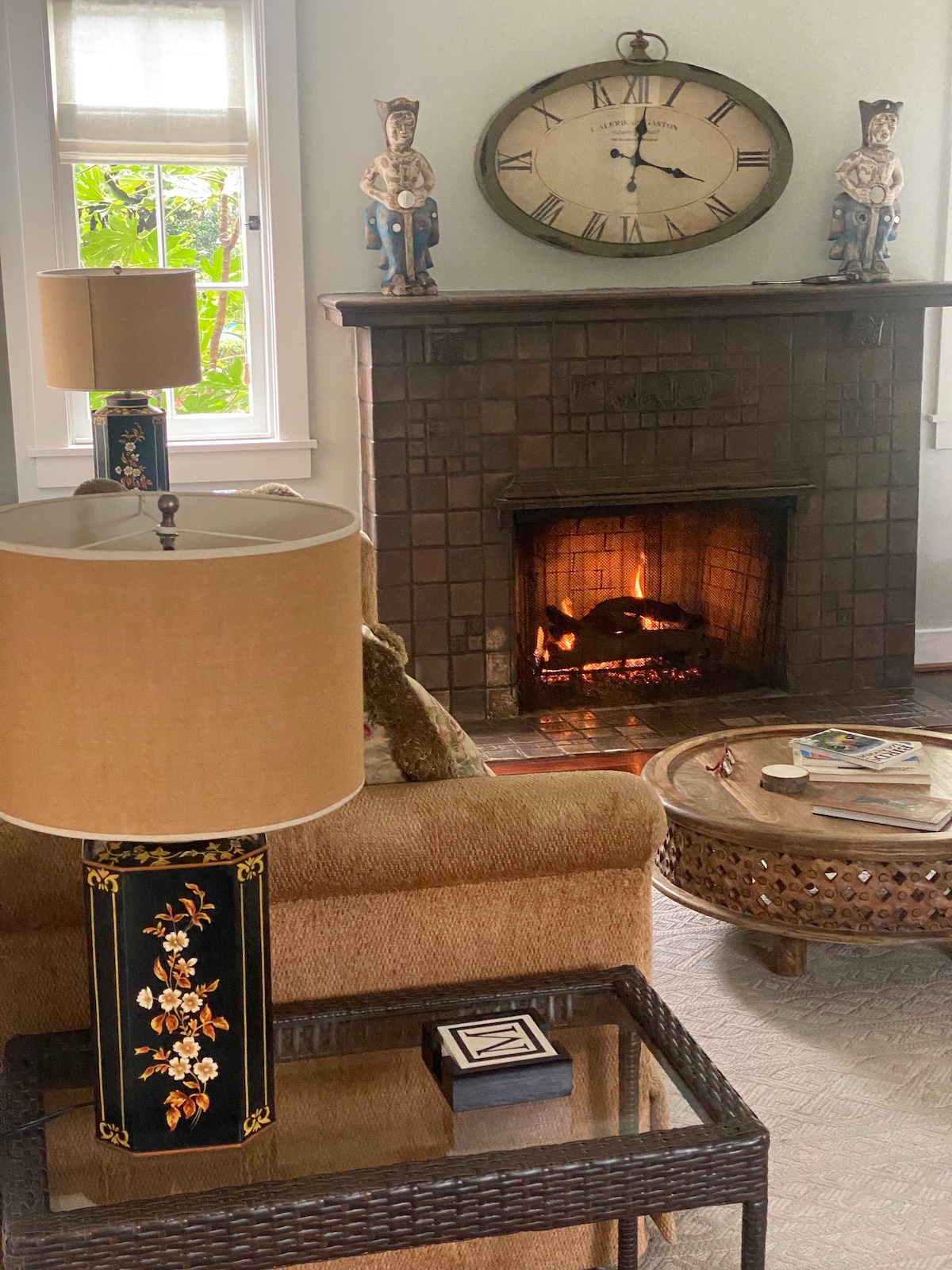 The room features a cozy seating area centered around a tiled fireplace, where a warm fire flickers. A decorative clock hangs above the mantel, flanked by two ornate figurines. A stylish table holds magazines and a small decorative item, accompanied by a lamp that provides soft lighting.