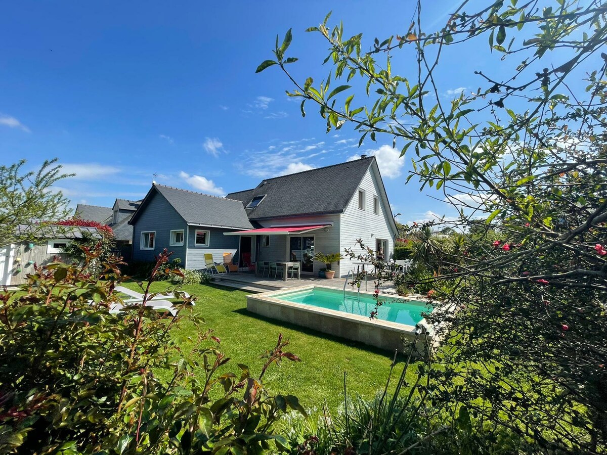 The exterior view of a wooden house is surrounded by lush greenery and colorful flowers. A well-maintained garden features lounge chairs and a small salt-water pool. The house includes a covered patio area with seating, under a clear blue sky.