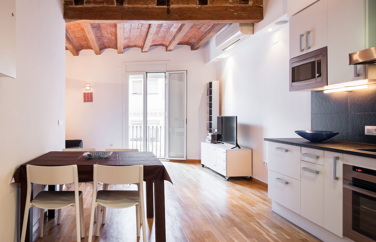 An open living area features a dining table with four chairs and a modern kitchen. A sleek entertainment unit holds a television, while large doors lead to a small balcony. Exposed wooden beams add character to the ceiling, complemented by warm hardwood flooring.