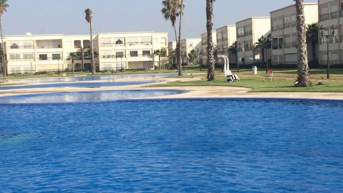 A series of inviting swimming pools are visible, framed by neat green lawns and palm trees. The water reflects bright blue tones under the clear sky. In the background, modern low-rise buildings provide a serene setting, enhancing the tranquil ambiance.