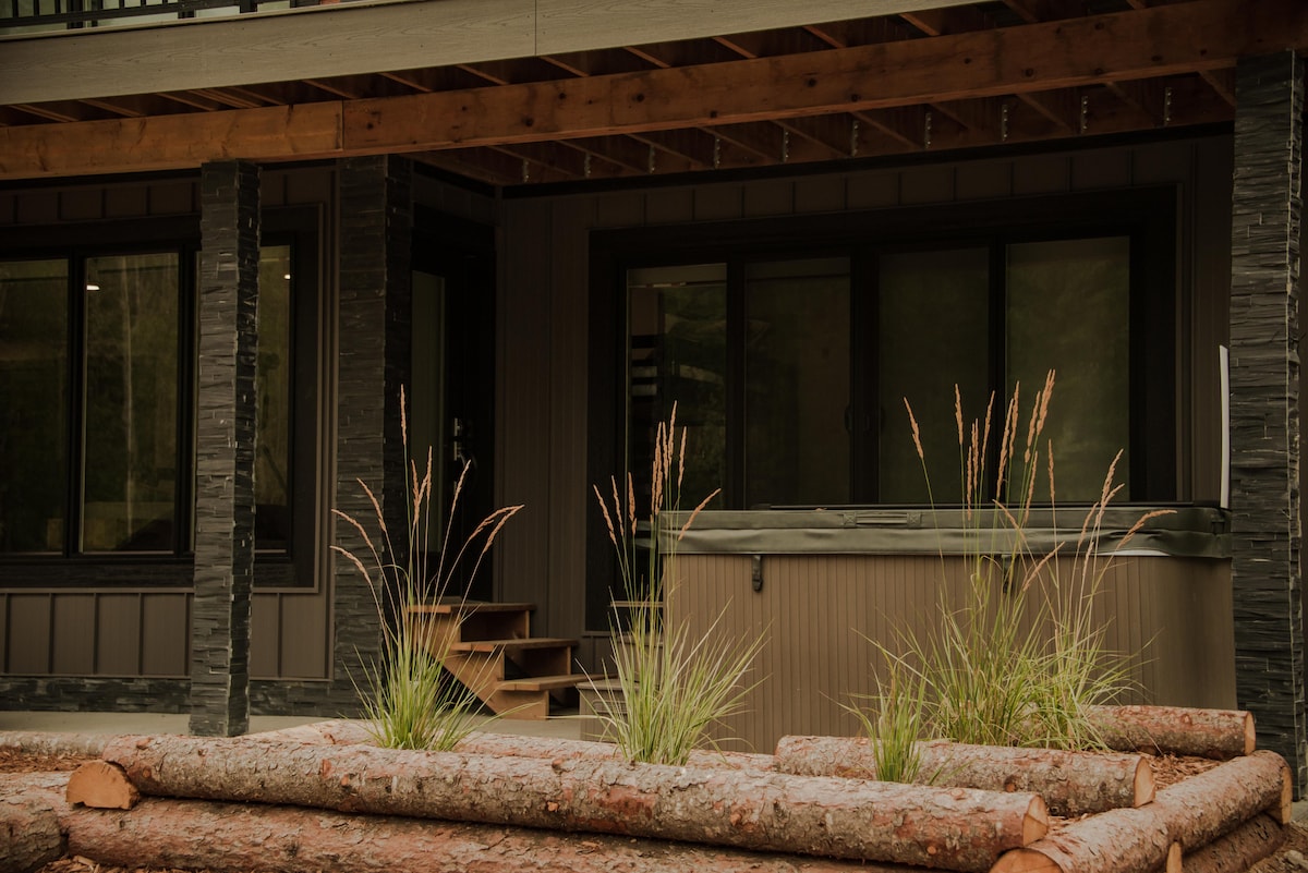 A concrete patio area is visible, featuring a hot tub surrounded by a wooden planter with tall grass. Steps lead up to the lodge, which has large windows and a covered entrance beneath a wooden overhang.