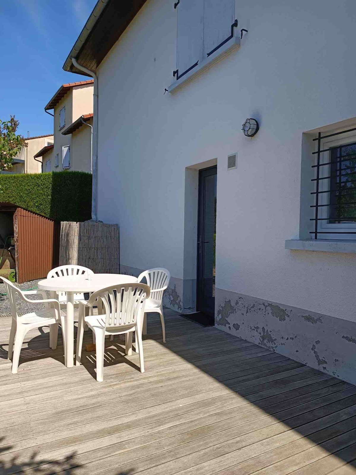 A small wooden terrace features a round table surrounded by four stacked white chairs. The entrance door is located beside a window, framed by a light exterior wall. Lush greenery is visible in the background, contributing to a tranquil outdoor space.