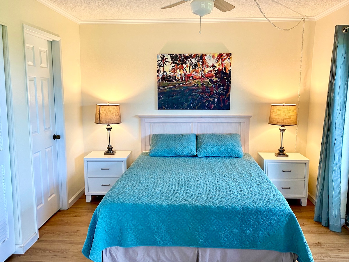 A cozy bedroom features a spacious bed dressed in a turquoise coverlet. Two bedside tables with lamps provide warm lighting, while a vibrant artwork hangs above the headboard. Light-colored walls and wooden flooring contribute to a serene atmosphere.