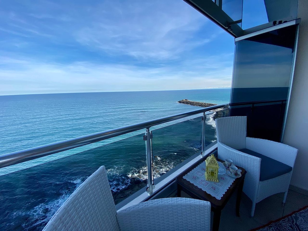 A balcony viewpoint captures a serene ocean scene, featuring a calm blue sea stretching towards the horizon. Two wicker chairs are set beside a small table, inviting relaxation. The glass railing offers unobstructed views of the gentle waves lapping at the nearby shore.