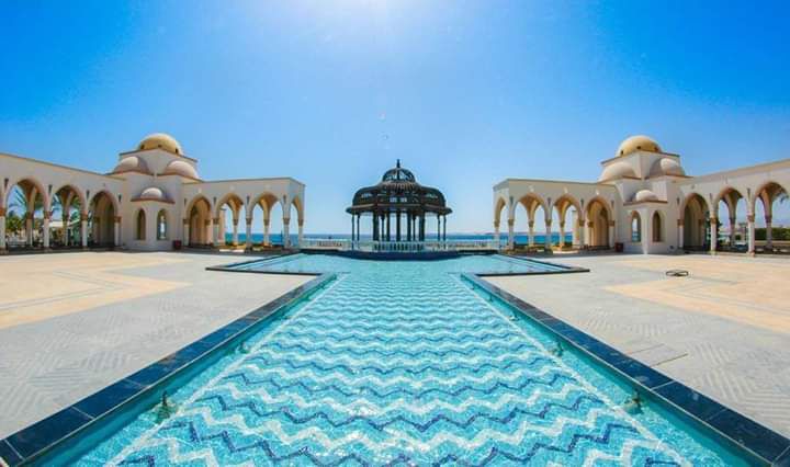 A spacious outdoor area features a geometric pool with blue and white tiles, leading towards a central pavilion. Arched structures flank the sides, under bright blue skies. The sea is visible in the background, adding to the serene atmosphere of the location.