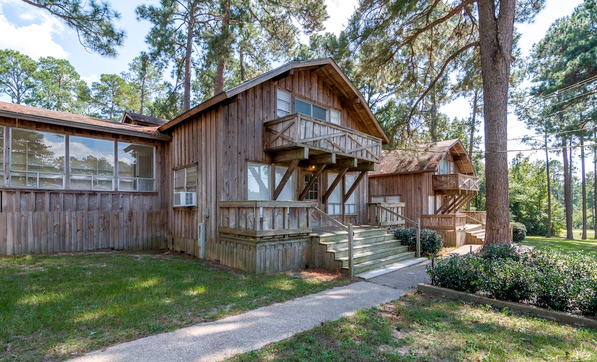 A rustic wooden cabin is set among tall pine trees, featuring a welcoming entrance with wooden steps and railings. The exterior showcases large windows that invite natural light, while the grassy area and well-maintained landscaping enhance the serene outdoor environment.