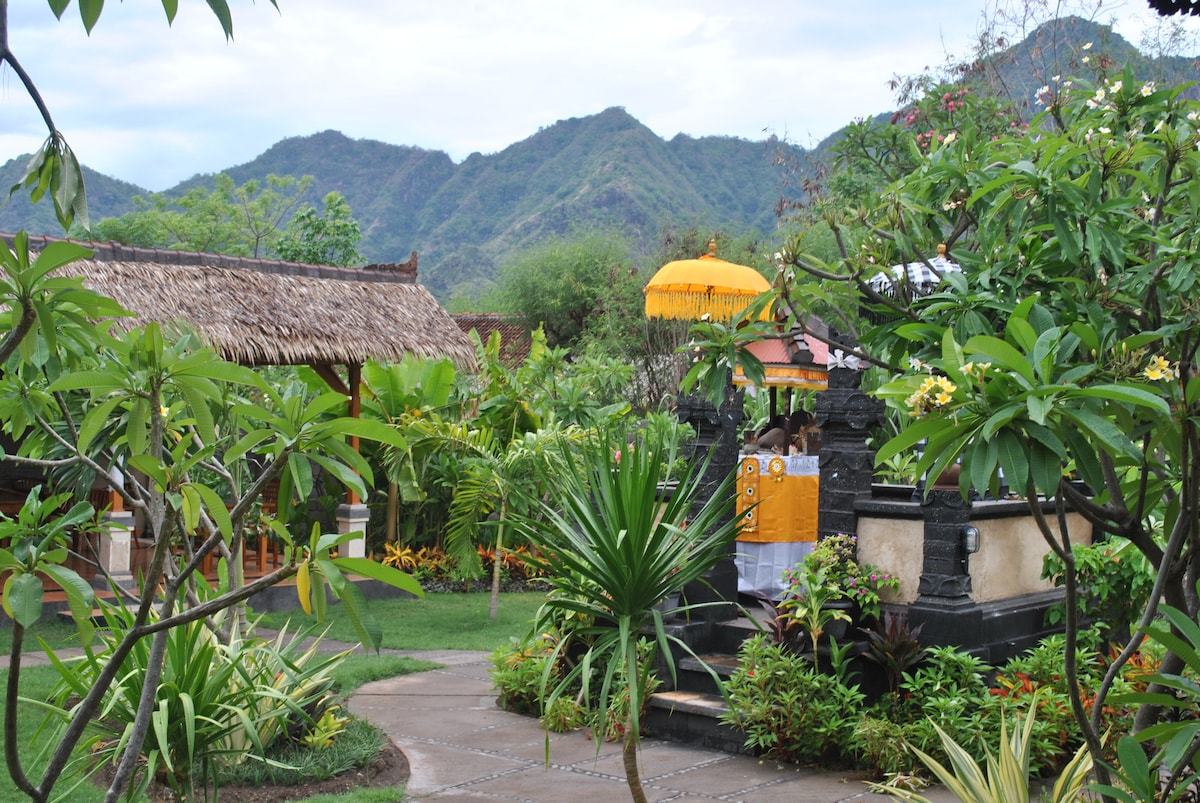 Lush tropical gardens are shown, featuring a thatched-roof gazebo surrounded by vibrant plants and flowers. A stone structure with decorative elements is visible alongside the mountain backdrop, conveying a serene environment. Pathways seamlessly connect different areas within the garden.