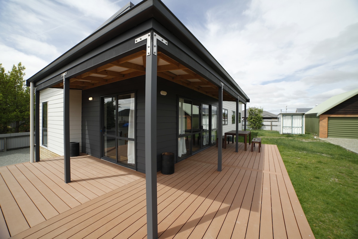 The exterior of a modern holiday home is depicted, featuring a spacious deck constructed with wooden planks. Large glass doors allow natural light to fill the interior, while a table and chairs sit on the deck, overlooking a grassy area.