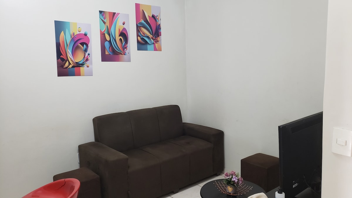 A living area is presented with a dark brown sofa complemented by two small seating cubes. Three colorful art pieces adorn the wall, adding visual interest. A flat-screen television is positioned to one side, while a small table is set with decorative items.