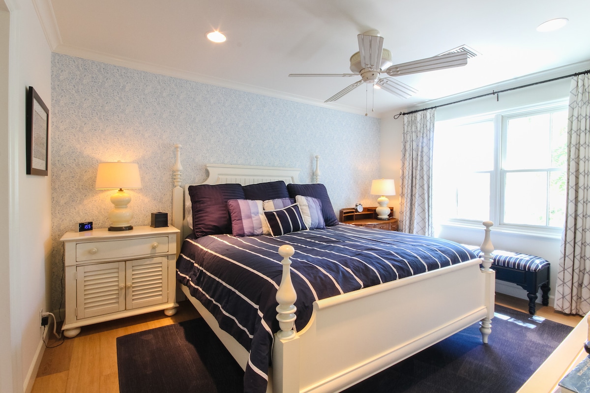 A serene bedroom is presented with a spacious white bed adorned in navy blue striped bedding. Soft lighting is provided by bedside lamps on white nightstands. Light streams through a window with sheer curtains, illuminating a cozy area featuring an upholstered bench at the foot of the bed.