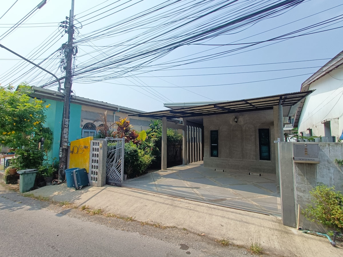 A well-kept exterior shows a spacious concrete driveway leading to a covered area. Lush greenery is visible along the perimeter, with vibrant plants adding color. Utility poles and overhead wires are present in the background, indicating the residential setting.