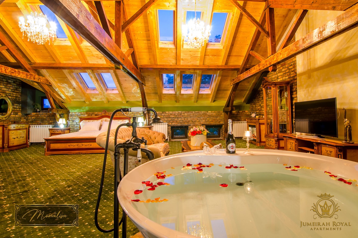 An inviting loft-style apartment features a spacious bathtub adorned with rose petals, set beside elegant wooden furnishings. Skylights provide natural light, illuminating the room's rich textures and calming colors. A luxurious king-sized bed is positioned nearby, enhancing the overall serene ambiance.