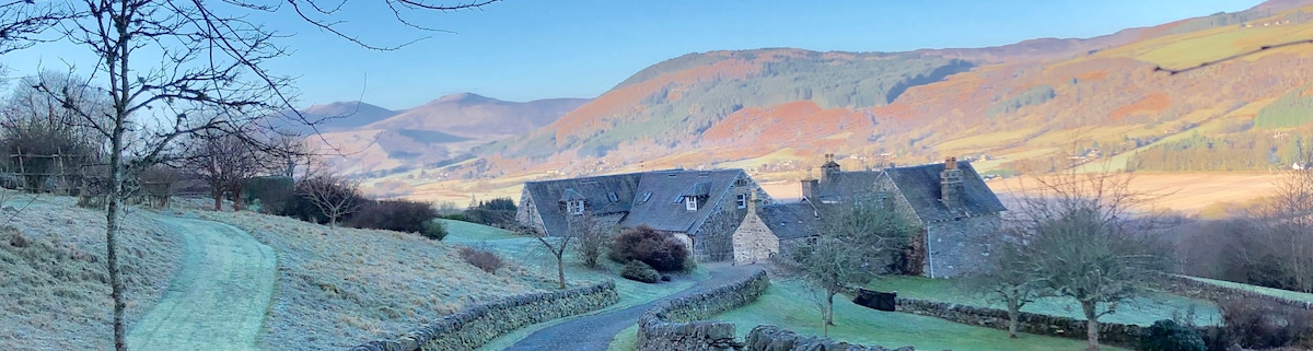 A scenic view of the property nestled in a rural landscape, surrounded by rolling hills. The stone buildings, with their traditional slate roofs, are complemented by frost-covered grounds and trees, reflecting the serene beauty of the Aberfeldy area.