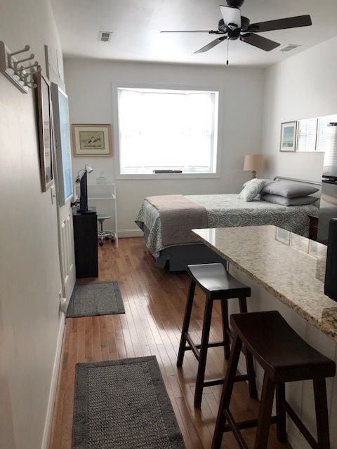 A studio apartment features a queen bed with a light-colored comforter, positioned under a window allowing natural light. A small dining area with two stools complements the kitchen, which is highlighted by granite countertops. Wooden floors enhance the warm ambiance, with a fan installed on the ceiling.