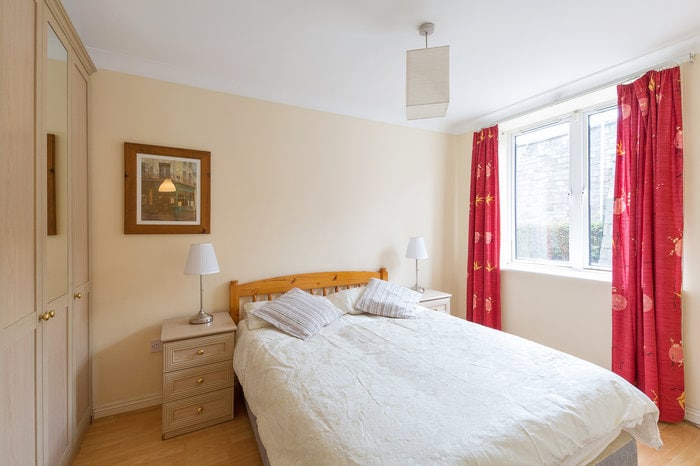A bright double room features a spacious bed with light bedding. Nightstands with lamps are positioned on either side of the bed. A framed artwork is displayed on the wall. Red curtains frame a window, allowing natural light to fill the room.