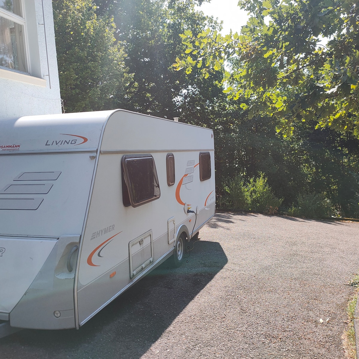A compact camper is parked on a gravel surface, featuring a smooth exterior design with subtle orange accents. Surrounding greenery and trees create a natural backdrop, providing shade and privacy. Sunlight filters through the leaves, casting gentle patterns on the ground.