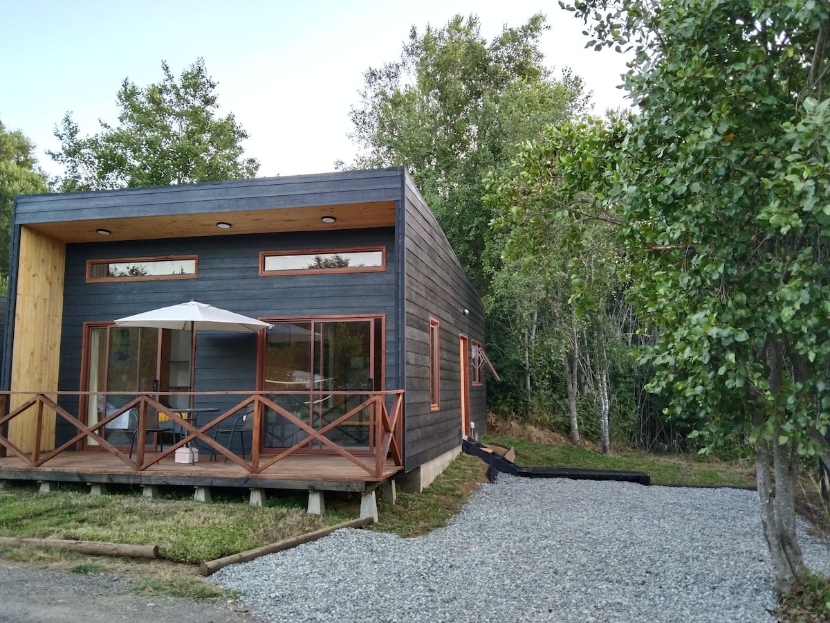 A modern cabin features a sleek wooden exterior, surrounded by trees. A shaded patio with a table and chairs is accessible through large windows. Gravel paths lead to the entrance, contributing to the natural ambiance of the setting.