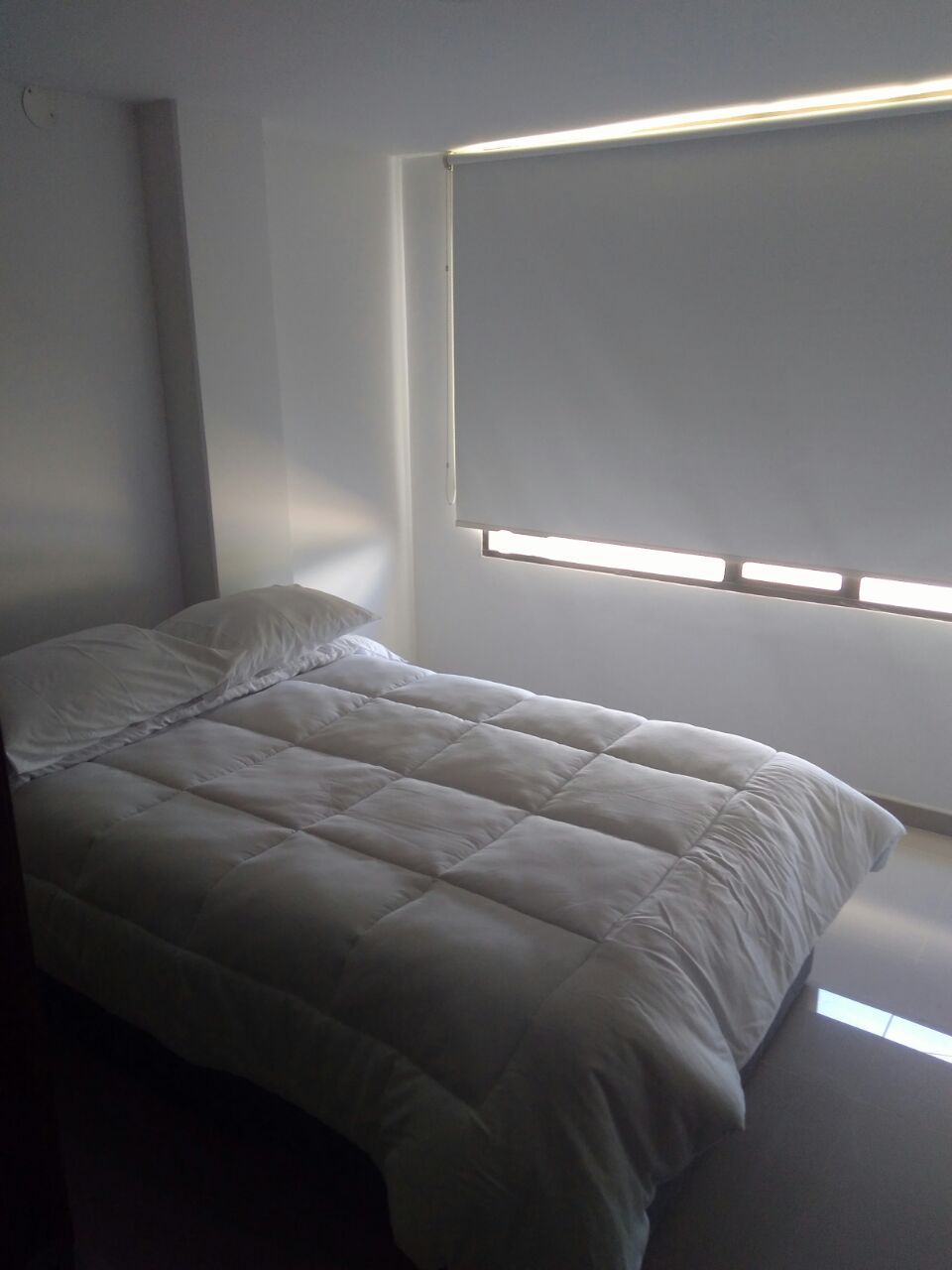 A well-lit sleeping area is presented with a spacious bed covered in a white quilt. Natural light filters through a large window fitted with a roller shade, creating a serene environment.