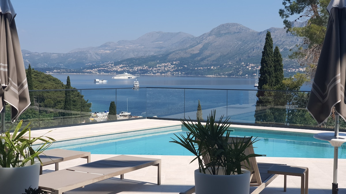 A tranquil outdoor area features a swimming pool with a view of the Adriatic Sea. Lounge chairs are positioned nearby, surrounded by greenery. Umbrellas offer shade against the sunlight, while distant mountains and a serene coastline form a picturesque backdrop.