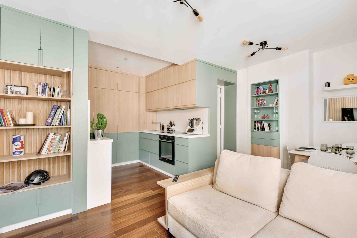 A modern open-plan living area showcases a light-colored sofa facing a well-equipped kitchen, featuring wood cabinetry and mint green accents. Bookshelves filled with books add character to the space, while contemporary lighting fixtures contribute to the overall bright and inviting ambiance.