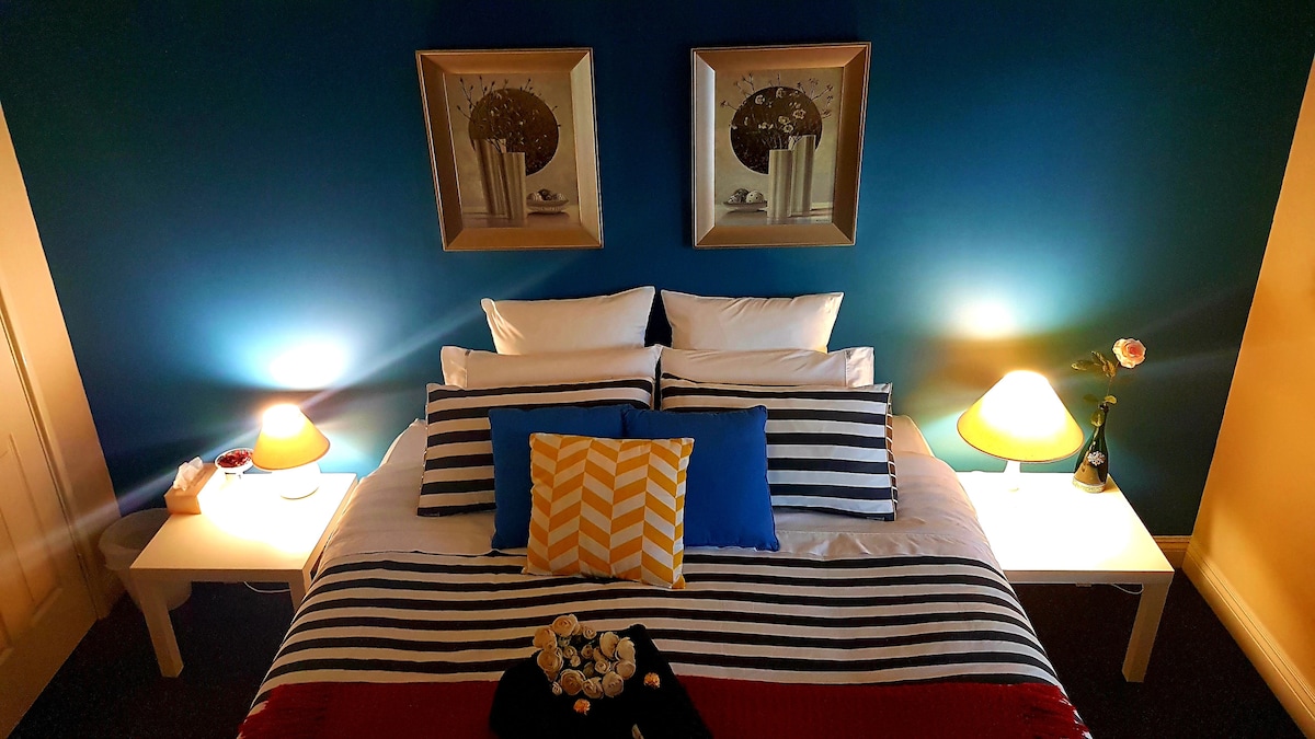 A spacious bed is adorned with striped bedding and colorful accent pillows, positioned against a deep blue wall. Two framed artworks hang above the bed, while bedside tables with lamps provide warm lighting. A touch of decor, such as a flower, adds a welcoming feel.