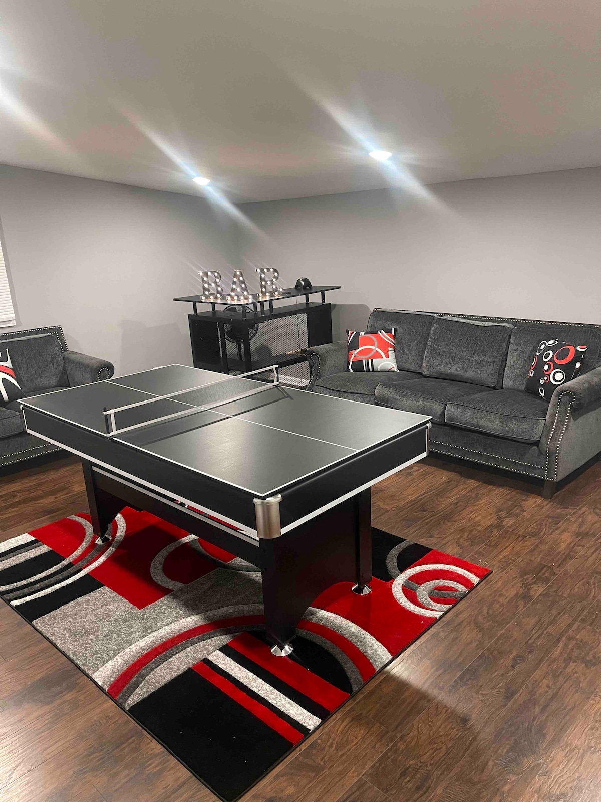 A recreational area features a modern ping pong table at its center, surrounded by comfortable seating options. Two gray sofas with decorative pillows provide a cozy atmosphere. The floor is covered with a patterned rug, complementing the neutral walls and bright overhead lighting.