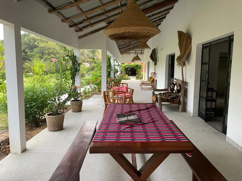 A spacious outdoor terrace is depicted, featuring a long wooden dining table set for meals. Surrounding greenery and potted plants provide a natural backdrop, while decorative light fixtures hang from the ceiling, enhancing the inviting atmosphere. Comfortable seating options are also presented along the terrace.