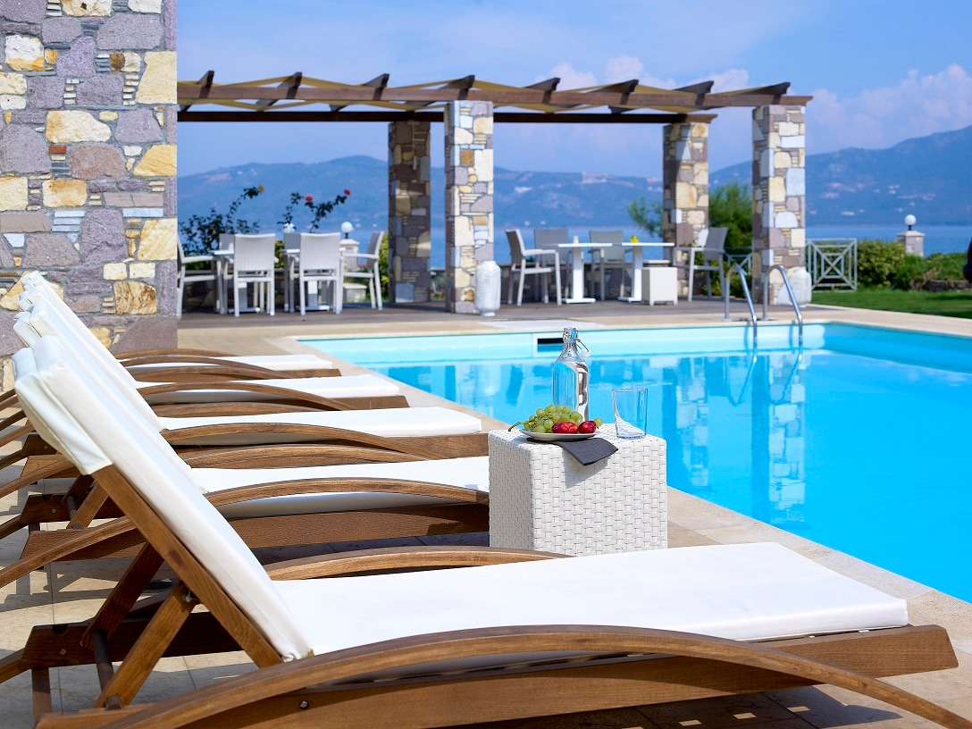 Lounge chairs with white cushions are aligned beside a sparkling pool, offering relaxation under the sun. A small table holds fresh fruit and drinks. In the background, a stone pavilion provides dining spaces overlooking the serene water and distant mountains.