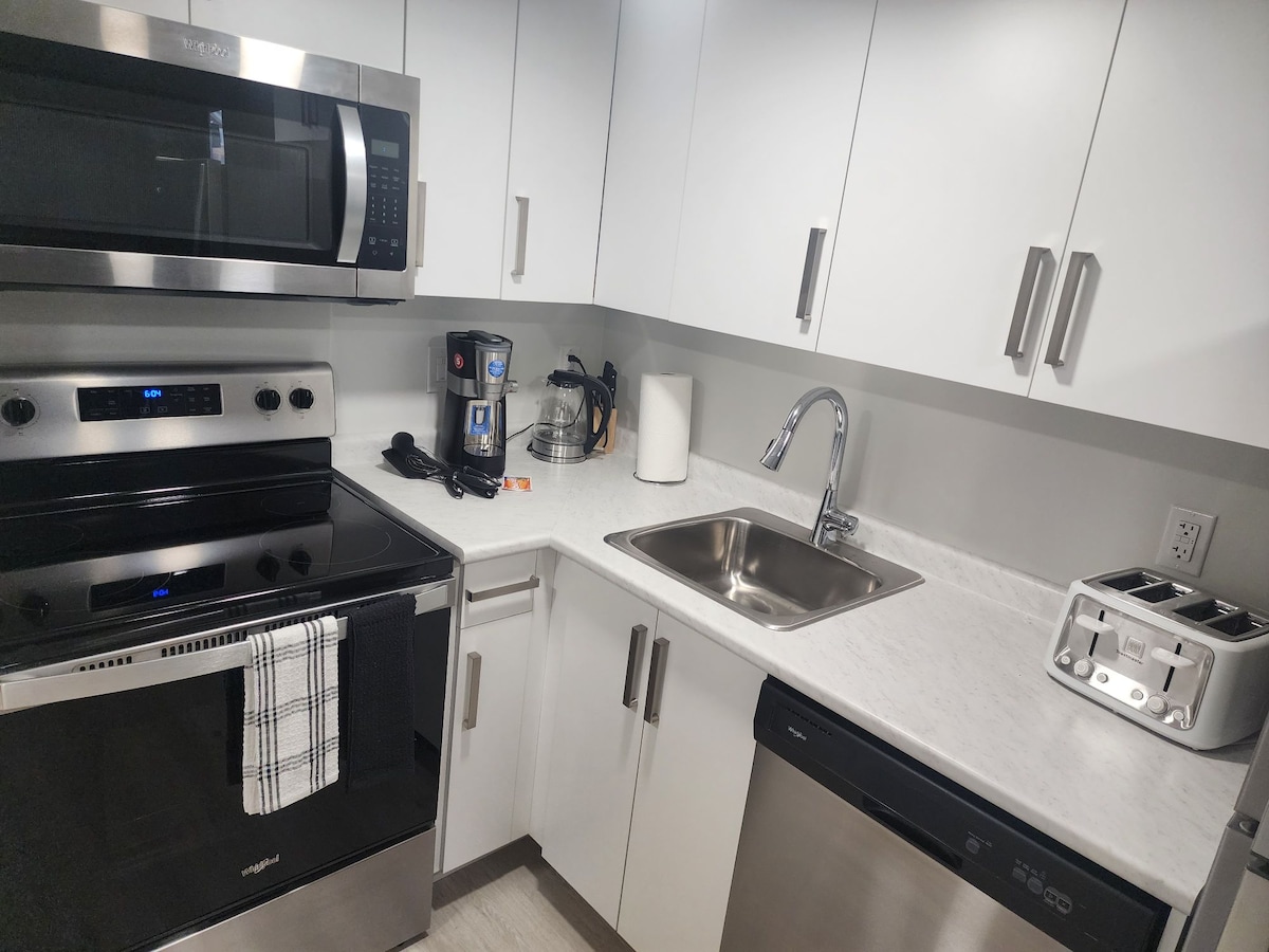 A modern kitchen setup features stainless steel appliances including a microwave, oven, and dishwasher. A sleek sink is paired with a stylish faucet, and a compact coffee maker is visible. The countertop is finished in light stone, complemented by crisp white cabinetry.