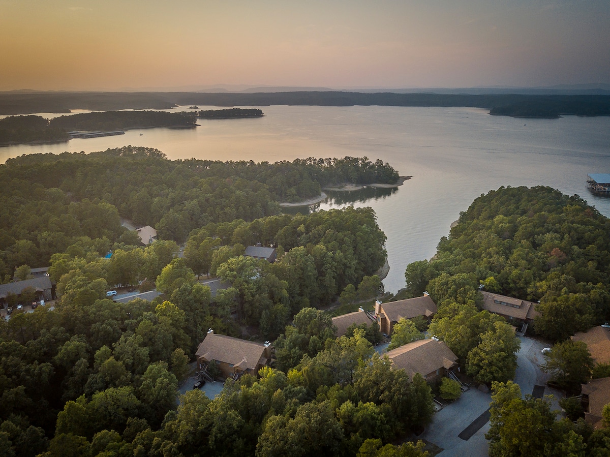 An aerial view captures the serene landscape of Lake Ouachita, surrounded by lush greenery and gentle hills. The expansive waters reflect the soft hues of sunset, creating a calming scene. Nearby structures can be seen nestled among the trees, highlighting the natural setting.