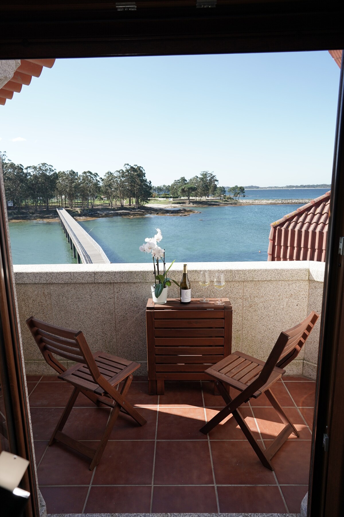 A cozy balcony setting features two wooden chairs beside a small table adorned with an arrangement of flowers and a wine bottle. A picturesque view of a blue waterway and a pedestrian bridge can be seen in the background, framed by trees lining the shore.