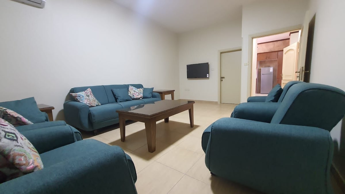 The living room features two comfortable sofas and matching armchairs arranged around a wooden coffee table. A mounted flat-screen TV is positioned on the wall. Light-colored walls and tiled flooring create a neutral backdrop for the cozy seating area.