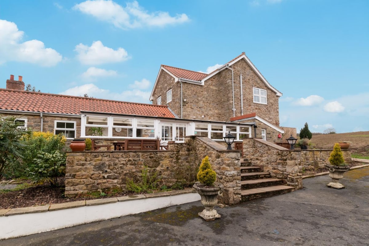 A stone cottage features a two-story design with a sloped roof and multiple windows. A spacious patio area with outdoor seating is surrounded by manicured gardens. Stone steps lead up to the entrance, framed by ornamental planters, against a clear blue sky.