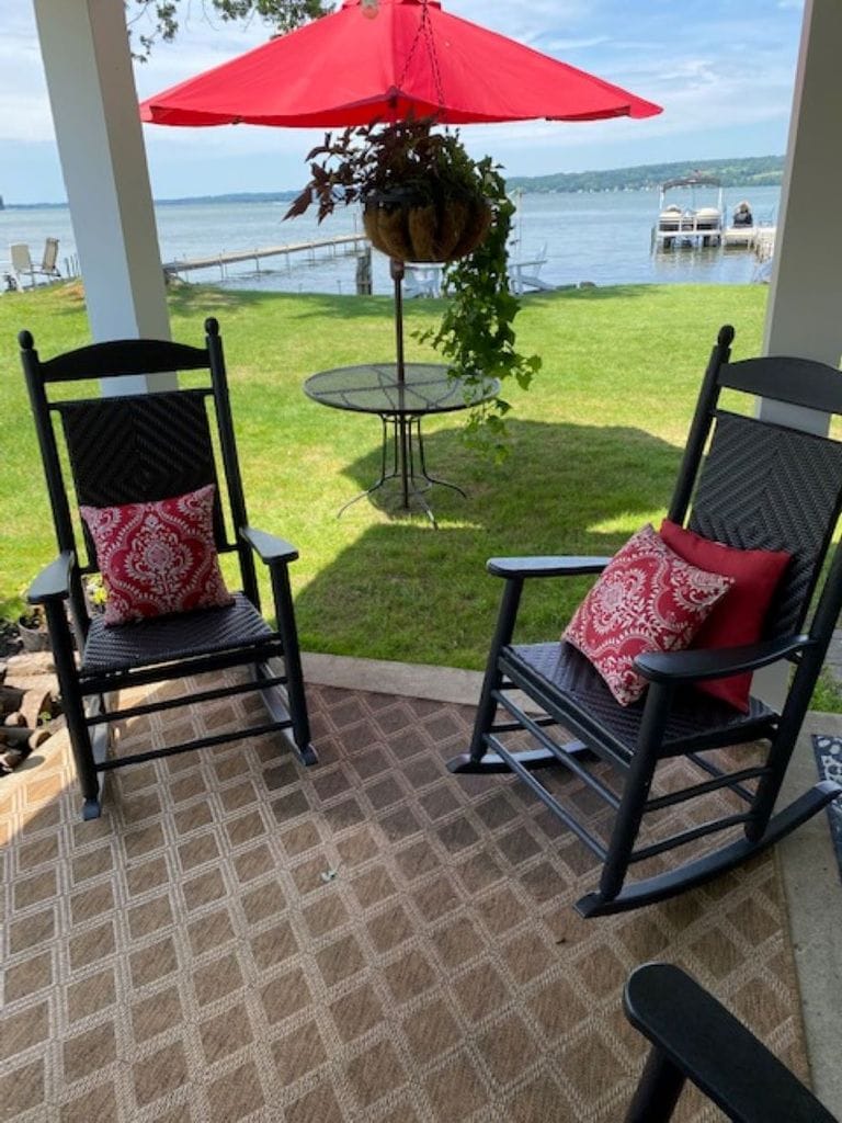 Two black rocking chairs, each adorned with red patterned cushions, are positioned on a textured area rug. A small round table stands nearby, with a hanging plant above. In the background, expansive green lawn and Chautauqua Lake can be seen under a clear sky.