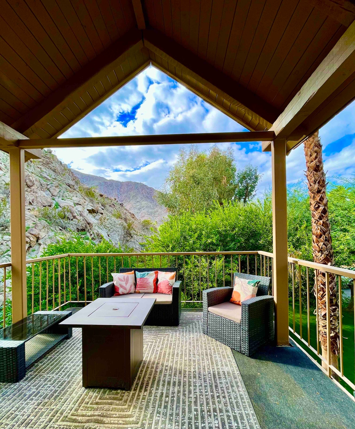 A private patio is framed by a wooden structure, featuring a natural view of mountains and greenery. Outdoor seating is offered with comfortable chairs and a central table, complemented by bright cushions. Light clouds drift across a clear blue sky, enhancing the serene atmosphere.