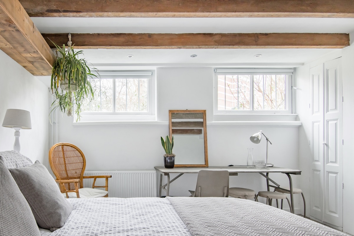 A serene bedroom is presented, featuring a spacious bed with a textured blanket. Large windows allow natural light to fill the space, complemented by wooden beams overhead. A desk with a chair and a decorative plant create an organized workspace, enhancing the room's functionality.