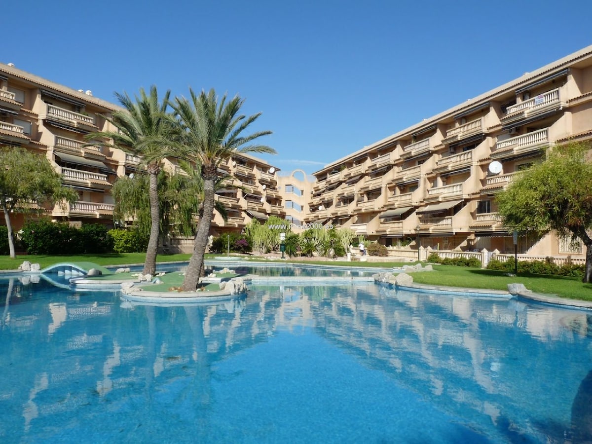 A tranquil communal pool is surrounded by well-maintained gardens, with palm trees positioned around its perimeter. The building features multiple levels, each with balconies overlooking the pool area. Clear blue skies reflect on the water, enhancing the relaxed setting.