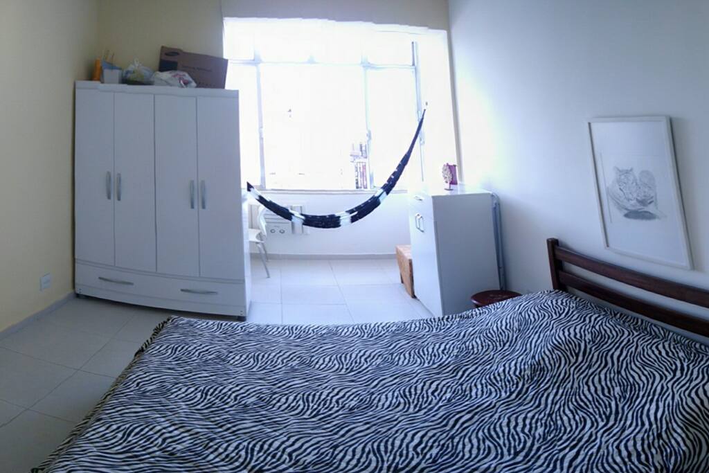 Quarto com janelão (muito claro! A luz está apagada), cama de casal, ar condicionado, ventilador de teto, armário 4 portas, mesa de cabeceira, mesinha de estudos com cadeira, rede e cômoda