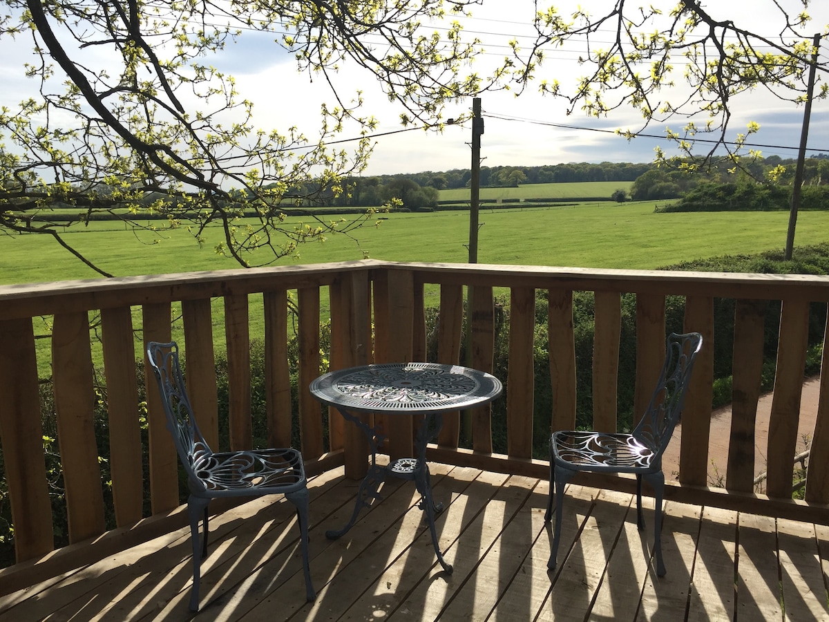 A raised deck features a round table and two ornate chairs, set against a backdrop of lush green fields and distant trees. Soft light filters through the leaves above, creating a serene outdoor space for relaxation.