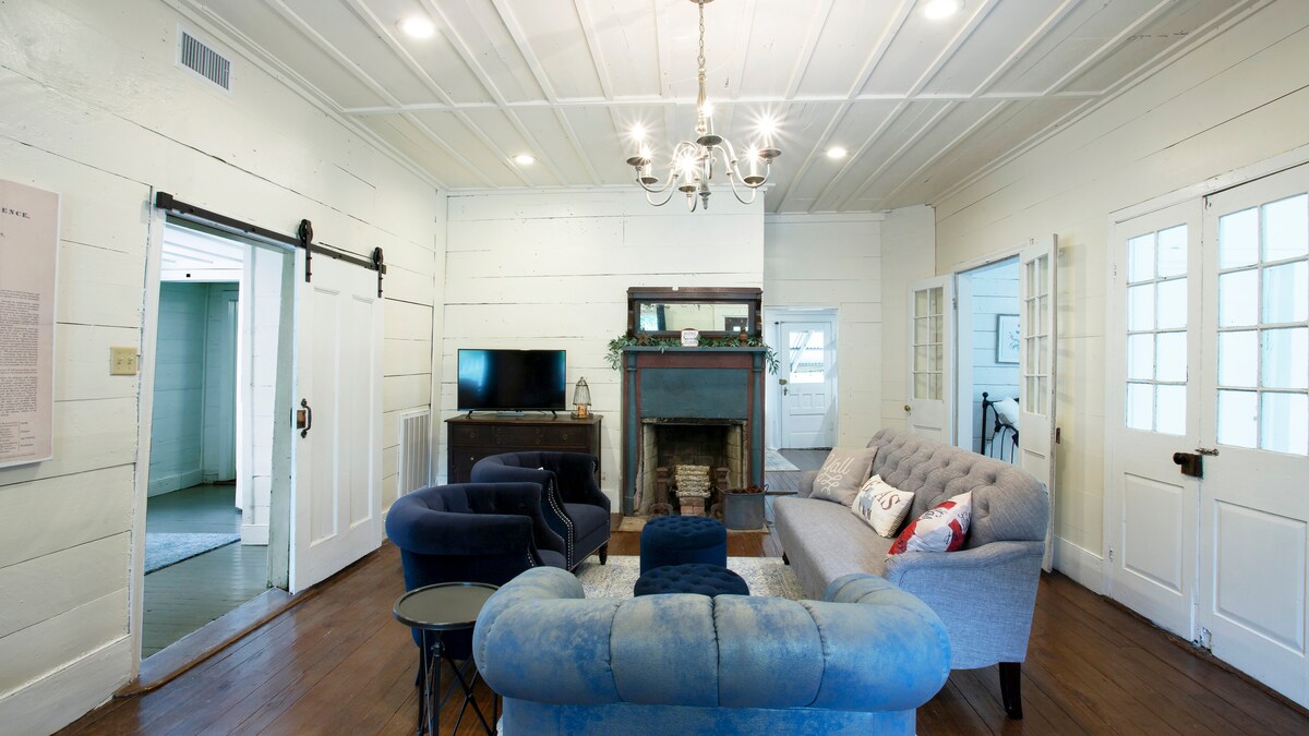 A spacious sitting area is present, featuring a plush sofa and a velvet chair. A vintage chandelier hangs from the ceiling, illuminating the space. A fireplace adds character, while natural light streams through multiple windows, enhancing the room's inviting atmosphere.