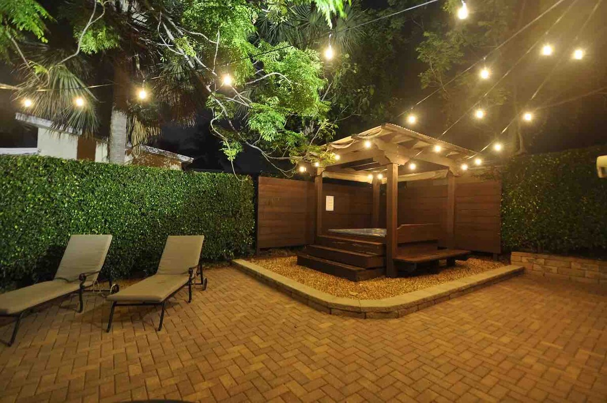 A landscaped backyard features a stylish hot tub under soft string lights, creating a cozy ambiance. Comfortable lounge chairs are positioned nearby on a paved patio, surrounded by lush greenery. The space is designed for relaxation and evening gatherings.