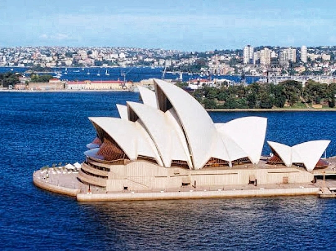 The Sydney Opera House is viewed from above, showcasing its distinctive sail-like structures against a backdrop of blue water. Buildings and greenery of the shoreline are visible in the distance, creating a coastal cityscape.